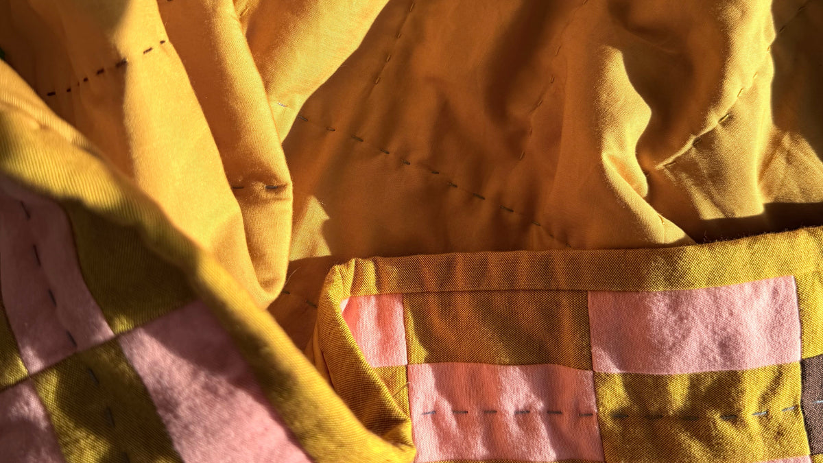 A close-up of a quilted fabric item, likely a blanket or quilt, featuring a patchwork design with mustard yellow, pink, and brown piecing under warm, direct sunlight.